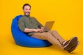 Portrait of attractive cheery man sitting in bag chair using laptop typing project isolated over bright yellow color Royalty Free Stock Photo