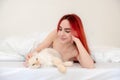 Portrait of an attractive, content, young, sexy red-haired woman lying relaxed in bed, enjoying and snuggling with a soft cat in Royalty Free Stock Photo
