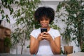 Portrait of attractive curly African young woman holding mobile phone in hands, enjoying web surfing, communicating in Royalty Free Stock Photo