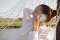 Portrait of attractive delighted woman wearing white shirt sitting in hammock on the bank of the river and talking smart phone Royalty Free Stock Photo