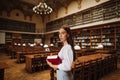 Portrait of attractive female student with book in hands at university library, looking at camera with serious face on background Royalty Free Stock Photo