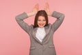 Portrait of attractive flirty young woman in business suit showing bunny ears over head and smiling, having fun