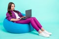 Portrait of attractive focused skilled cheerful girl lying in bag chair using laptop isolated over bright teal color Royalty Free Stock Photo