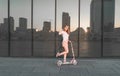 Portrait of attractive girl riding on pink scooter against background of dark glass wall, reflection of sun and city landscape Royalty Free Stock Photo