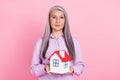 Portrait of attractive gray-haired woman holding in hands house mini decor over pink pastel color background