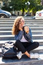 Portrait of an attractive happy woman talking on the phone holding coffee cup while sitting on stairs at the city street Royalty Free Stock Photo