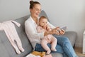 Portrait of attractive happy woman with bun hairstyle wearing white shirt and jeans sitting on sofa with baby daughter and Royalty Free Stock Photo