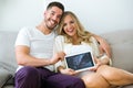 Attractive happy young man and pregnant smiling woman looking at camera while holding digital tablet at home.