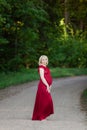 Beautiful pregnant woman in stylish long red maternity dress looking dreamy on lonely road in forest Royalty Free Stock Photo