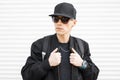 Portrait of an attractive man hipster in a black cap in stylish black sunglasses in a trendy jacket with a sports backpack Royalty Free Stock Photo