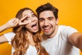 Portrait of attractive man and woman taking selfie photo and showing peace sign, isolated over yellow background Royalty Free Stock Photo