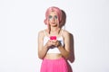 Portrait of attractive party girl in pink wig, with bright makeup, holding smartphone and looking at camera confident