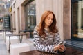 Portrait of attractive redhead young woman sitting outdoors cafeteria using smartphone chatting with friends looking to Royalty Free Stock Photo