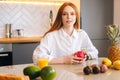 Portrait of attractive redhead young woman sitting at table with exotic tropical fruit in kitchen room with light modern Royalty Free Stock Photo