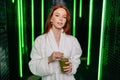 Portrait of attractive redhead young woman wearing white bathrobe, holding glass with freshly squeezed vitamin juice in Royalty Free Stock Photo