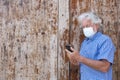 Portrait of attractive senior man with white hair wearing face mask due to coronavirus using smart phone standing against a wooden Royalty Free Stock Photo