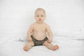 Portrait of attractive serious grey-eyed plump cherubic baby infant toddler wearing grey pants sitting on white bed.