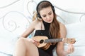 Portrait of Attractive Sexy Woman is Relaxing on Bedroom While Playing Ukulele Instrument and Listen Music on Headphones on Royalty Free Stock Photo
