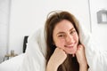 Portrait of attractive smiling woman relaxing in bed, lying covered in white sheets and warm duvet in morning, looking Royalty Free Stock Photo