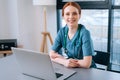 Portrait of attractive smiling young female doctor in blue green medical uniform sitting at desk with laptop on Royalty Free Stock Photo