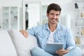 Portrait of an attractive smiling young man wearing casual clothes sitting on a couch at the living room, using digital tablet Royalty Free Stock Photo