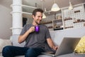 Smiling young man wearing casual clothes sitting on a couch in the living room, using laptop computer Royalty Free Stock Photo