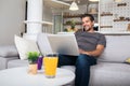 Smiling young man wearing casual clothes sitting on a couch in the living room, using laptop computer Royalty Free Stock Photo