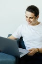 Portrait of an attractive smiling young man wearing casual clothes sitting on a couch at the living room, using laptop Royalty Free Stock Photo