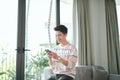 Portrait of an attractive smiling young man wearing casual clothes sitting on a couch at the living room, using mobile phone Royalty Free Stock Photo