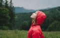 Portrait of attractive tourist girl in red raincoat stands on valley in mountains and looks up at cloudy sky. Pretty woman in Royalty Free Stock Photo