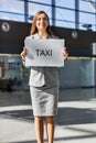 Portrait of attractive woman standing while holding white board with TAXI signage in arrival area at airport Royalty Free Stock Photo