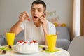 Portrait of attractive young adult man wearing white casual style T-shirt sitting at table and eating cake, talking smart phone Royalty Free Stock Photo