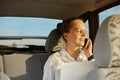 Portrait of attractive young adult woman in optical glasses sitting in the car and talking on the cell phone, holding laptop, Royalty Free Stock Photo