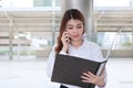 Portrait of attractive young Asian secretary woman talking on phone and looking paperwork in document folder at sidewalk of office Royalty Free Stock Photo