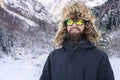 Portrait of an attractive young bearded man in a large fur hat and sunglasses against the snow-covered forest in the