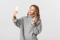 Portrait of attractive young blond girl in grey sweater holding smartphone, showing peace sign and taking selfie Royalty Free Stock Photo