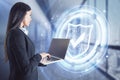 Portrait of attractive young european businesswoman with laptop standing in blurry office interior with glowing shield hologram.