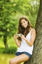 Portrait of attractive young smiling brunette girl wearing white shirt and making photos at summer green park with compact camera. Royalty Free Stock Photo