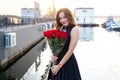 Portrait of attractive young woman with brown hair in little black dress smiling and holding a bouquet of red roses at sunset Royalty Free Stock Photo