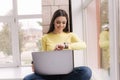 Portrait of attractive young woman using laptop in front of big window. Brunette woman in yellow sweatshirt working at Royalty Free Stock Photo