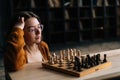 Portrait of attractive young woman wearing elegant eyeglasses thinking about chess move while sitting on wooden floor in Royalty Free Stock Photo