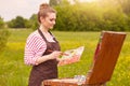 Portrait of attractive young woman wearing white shirt with red stripes and brown apron, holding palette of colours and brush, Royalty Free Stock Photo