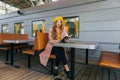 Portrait of awesome girl, wearing coat and yellow beret. Sitting on the train station read a book and looking straight. Autumn