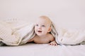 Portrait of baby hiding under the white blanket. Cute 8 month baby girl crawling in bed. Tummy Time. Textile and bedding for kids