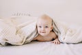 Portrait of baby hiding under the white blanket. Cute 8 month baby girl crawling in bed. Tummy Time. Textile and bedding for kids