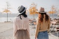 Portrait from back of brunette girl in hat talking with friend on nature background. Ginger lady in beige jacket