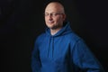 Portrait of bald and clean shaven man with glasses dressed in blue hoodie on a black background.