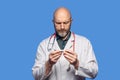 Portrait of bald doctor on blue background. Male health care worker in 40s, with grey beard holding white digital thermometer, Royalty Free Stock Photo