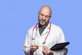 Portrait of bald doctor on blue background. Male health care worker in 40s, with grey beard holding white digital thermometer, Royalty Free Stock Photo