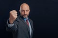 Portrait of bald man in business suit on dark background. Gentleman in his 40s with gray beard pointing clinched fist at viewer,